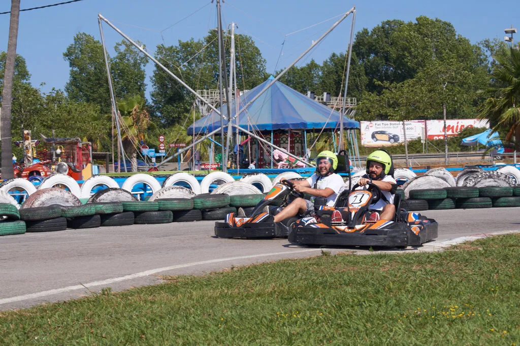 Dos adultos muy amigos compitiendo en los karts grandes. Están en medio de la curva 3. En el circuito de karts para adultos. Es un buen dia de fin de semana. En el background se ven las atracciones infantiles y personas disfrutando del dia.